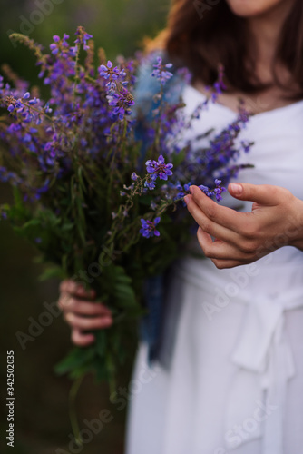 a girl in white dress holds wild flowers in her hands. the girl touches a bunch of purple flowers with her fingertips