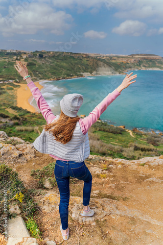 the girl stands on the mountain with her hands raised. view from the mountain to the sea. a girl in a pink jacket and jeans