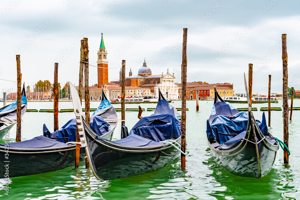 Venice, Italy. Empty Gondolas/ Gondole docked by lagoon mooring poles. Famous romantic city tour boat ride for tourists/ couples/ people. Bell Tower San Giorgio Maggiore Basilica Church in background