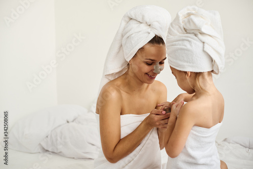 A young caucasian mother and little daughter with wrapped hair in white bath towels apply a clay mask on the mother faces