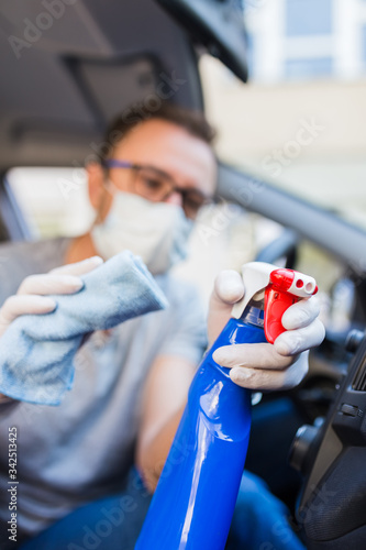 Man in face mask using disinfectant and microfiber cloth to clean car interior.