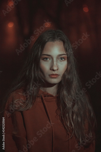 Portrait of a girl with long hair in a hoodie surrounded by darkness and red lights at night