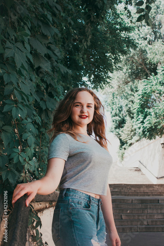 Young woman stands near a wall covered in green plants during the day while wearing a gray shirt and ripped jeans