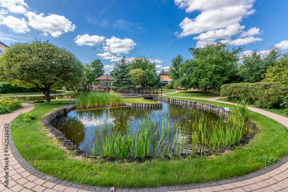 Fototapeta premium A park site with a little pond, surrounded by trees. A wooden bridge and gazebo in the background
