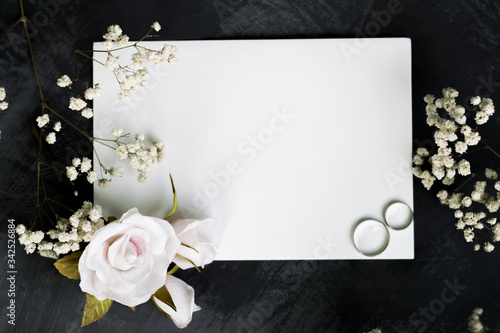 Gypsophila branches, roses and white gold wedding rings for a card or wedding invitation. Blank paper mock-up in minimalist style. White sheet of paper for inscription on a gray background, copyspace