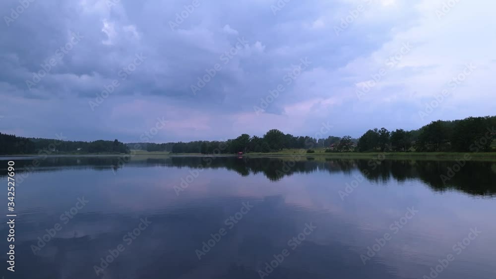 Slow motion. Gorgeous view of sunset on calm summer evening. Lake shore with green trees and plants reflecting in crystal clean mirror water surface. Sky covered with heavy thunder clouds. Slow motion. Gorgeous view of sunset on calm summer evening. Lake shore with green trees and plants reflecting in crystal clean mirror water surface. Sky covered with heavy thunder clouds.