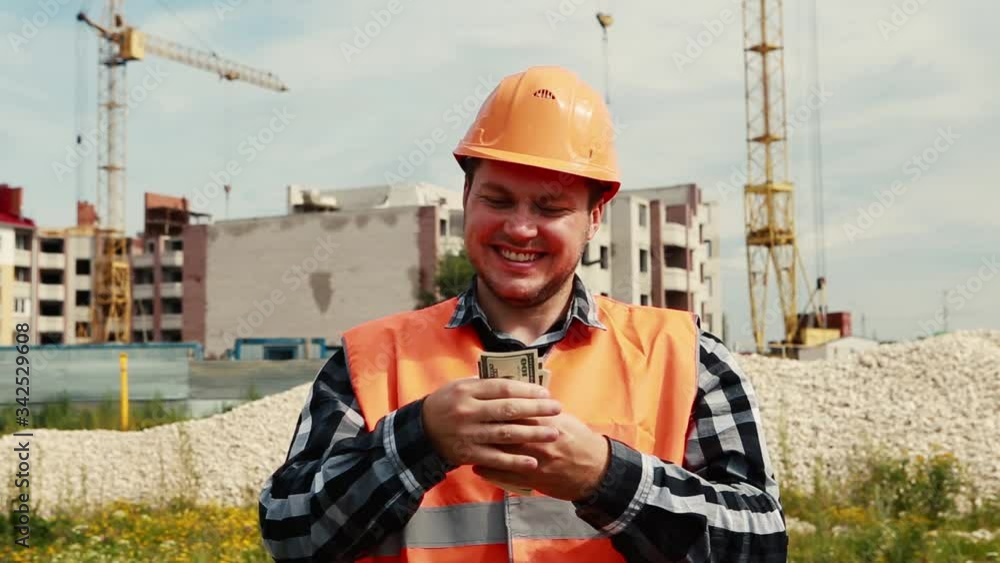 The builder counts the money against the background of the construction site and puts it in an orange helmet The builder counts the money against the background of the construction site and puts it in an orange helmet