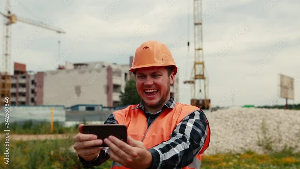 A man in an orange vest and orange helmet takes pictures of himself on a construction site on a mobile phone. A man in an orange vest and orange helmet takes pictures of himself on a construction site on a mobile phone.