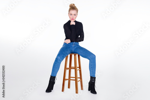 Sexy girl in tight jeans sitting on stool with legs apart and chin resting on fist.