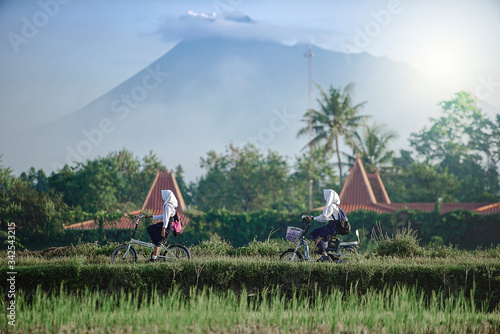Yogyakarta Indonesia June 28, 2020 : Students on the way go to school in Sleman village, with bycyle in the morning blue sky and merapi mountain view photo