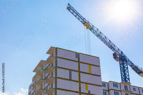 Construction crane on the construction of a residential building with a ventilated facade in the sun. The building has windows, a modern facade, no roof.