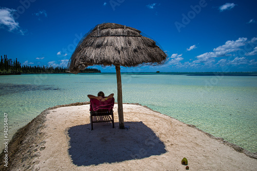 Фотография A woman relaxing on a beautiful beach under a sunshade overlooking a pristine crystal clear sea with some pines on the distance in the Ile of Pines, New Caledonia
