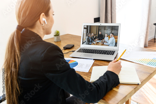 A woman in smart casual has online meeting from home, group of her colleagues on laptop screen. Back view