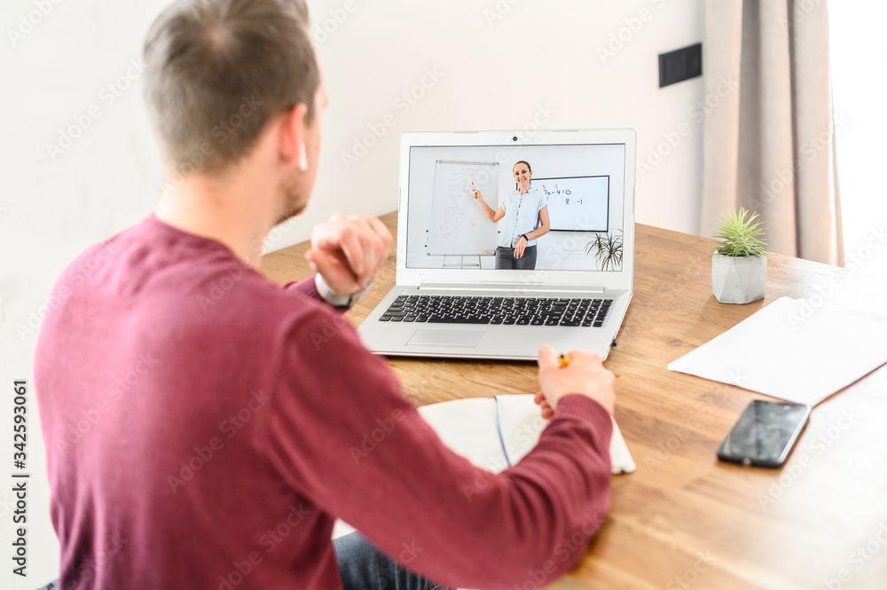 A young guy is studying online, watching webinars and writing in notebook, female teacher on the screen. Side view A young guy is studying online, watching webinars and writing in notebook, female teacher on the screen. Side view