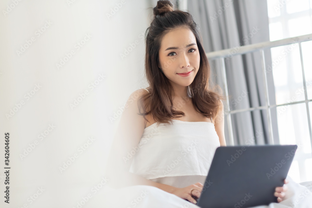 Pretty woman sitting in bed and smiling and looking at camera on morning. Positive Asian beautiful girl using laptop computer in white bedroom at home on holiday. Pretty woman sitting in bed and smiling and looking at camera on morning. Positive Asian beautiful girl using laptop computer in white bedroom at home on holiday.