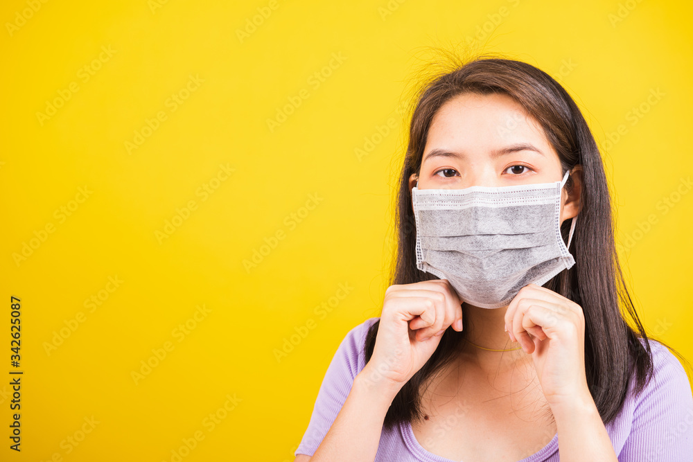 Closeup of portrait Asian young teen woman wearing face mask protective against coronavirus or COVID-19 virus showing demonstrating correct step, studio shot isolated yellow background Closeup of portrait Asian young teen woman wearing face mask protective against coronavirus or COVID-19 virus showing demonstrating correct step, studio shot isolated yellow background