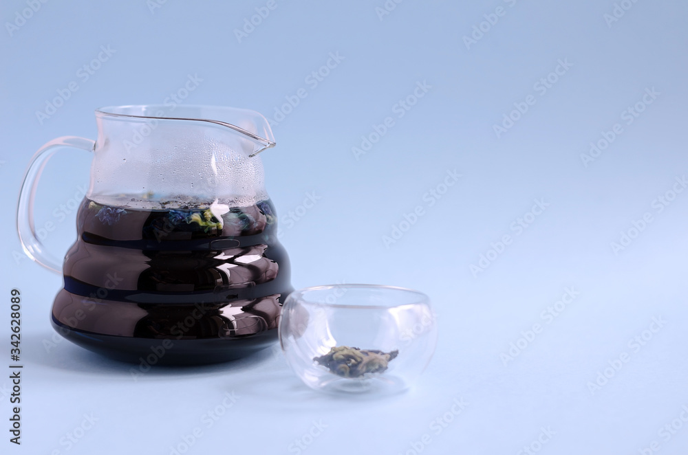 A glass teapot for brewing with blue tea stands on a blue background. Nearby is a small empty bowl. Space for text A glass teapot for brewing with blue tea stands on a blue background. Nearby is a small empty bowl. Space for text