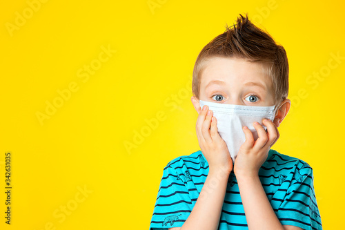 Portrait of a blue-eyed five-year-old European boy in a medical mask on a yellow background. The child put his hands to his face and stared. Fighting coronovirus, covid-19. Virus prevention.