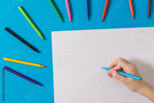 A hand with a blue marker and a white sheet of paper and a set of colored markers.