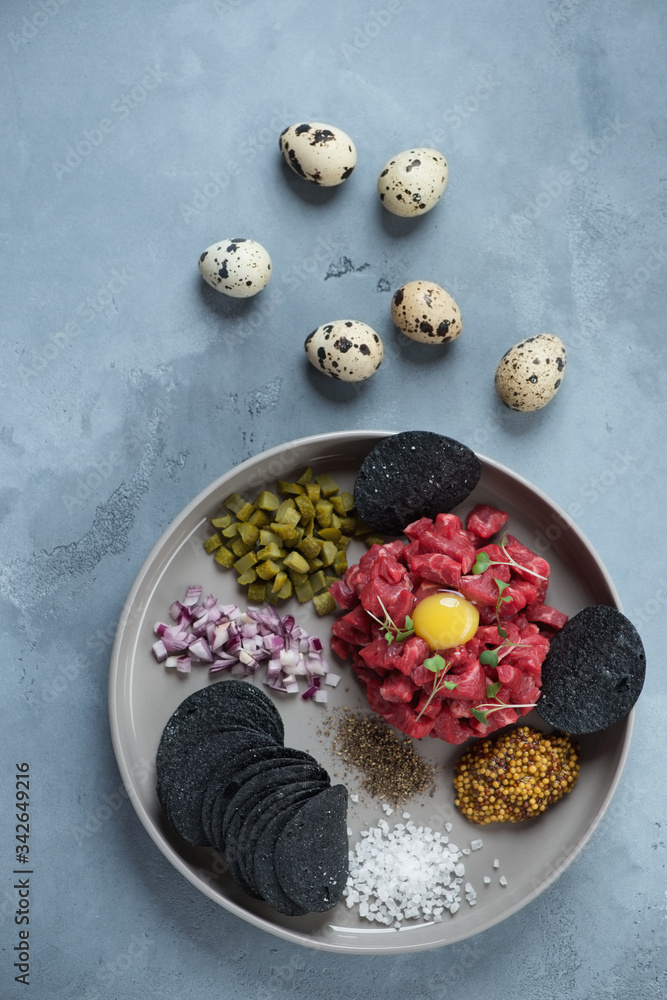 Round grey plate with beefsteak tartare, vertical shot on a grey concrete background, above view Round grey plate with beefsteak tartare, vertical shot on a grey concrete background, above view