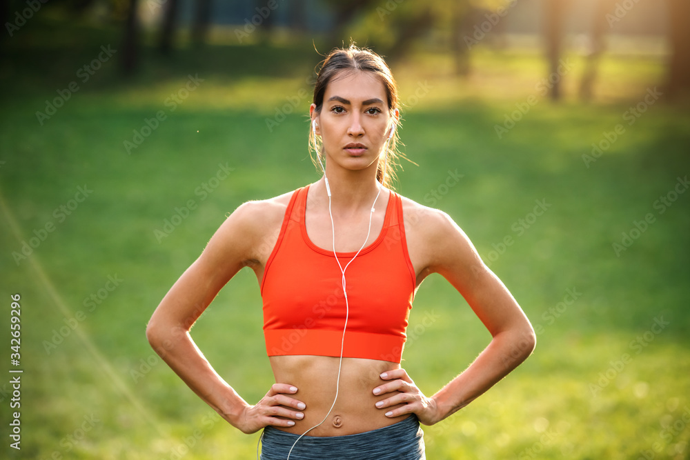 Portrait of dedicated attractive slim young sportswoman having earphones in ears and standing in nature with hands on hips.