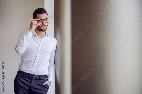 Young geeky thoughtful businessman in shirt leaning on pillar and gazing at something.