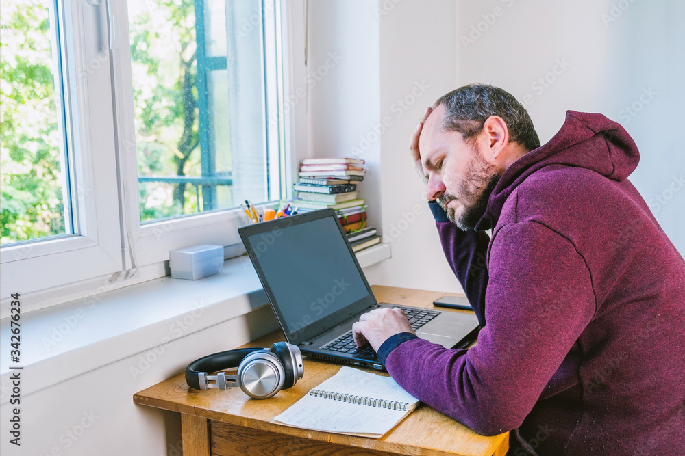 Man in hoodie sitting behind the laptop next to window, eyes closed, unsuccessful, tired, home office work concept Man in hoodie sitting behind the laptop next to window, eyes closed, unsuccessful, tired, home office work concept