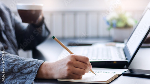 Close-up of hand businesswoman writing on notebook to planing her strategy while she working with laptop and drink coffee