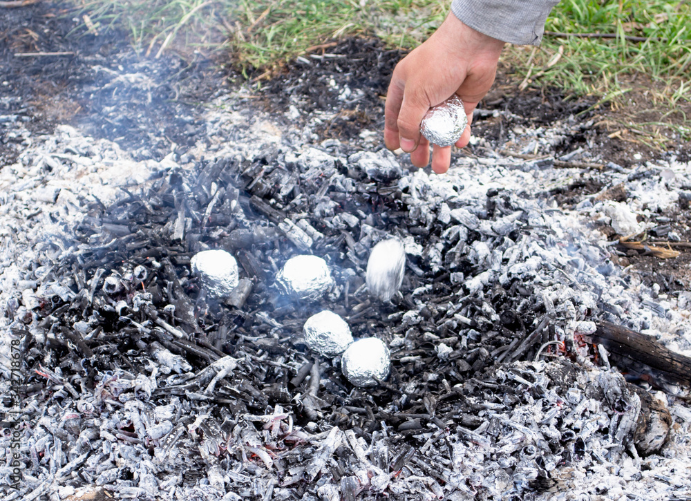 Potato wrapped in foil is cooked on hot coals. Hand throws potatoes in a bonfire. Potato wrapped in foil is cooked on hot coals. Hand throws potatoes in a bonfire.