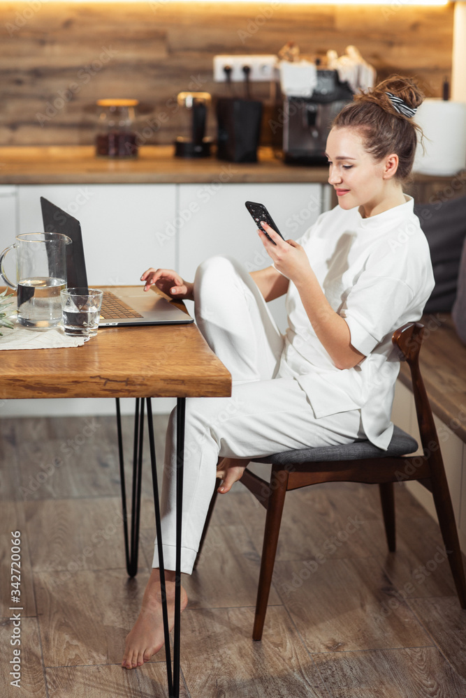 Young woman sits at the kitchen table using a laptop and talking on a cell phone and smiling. Successful girl laughing and working at home. Beautiful stylish woman smiling and relaxing at home.