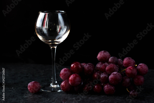 a glass glass stands on the table against a black background. There are red grapes nearby. On top of the glass is poured wine from a bottle with a dark glass