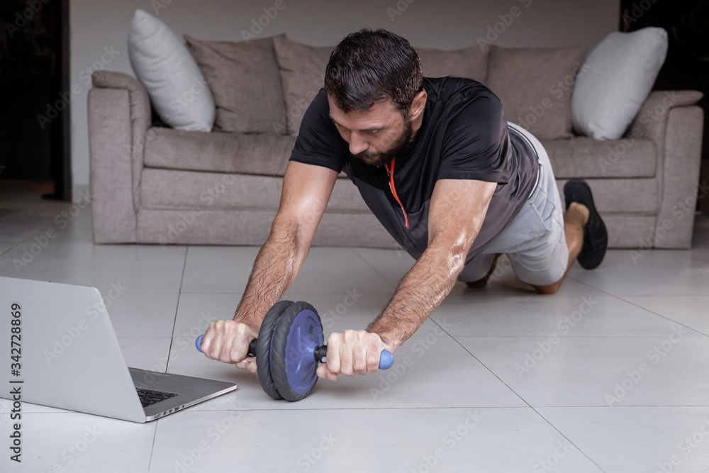 Obraz premium Young man exercising and working out online with laptop or phone at home during covid-19 pandemic quarantine. Sports and healthy lifestyle concept