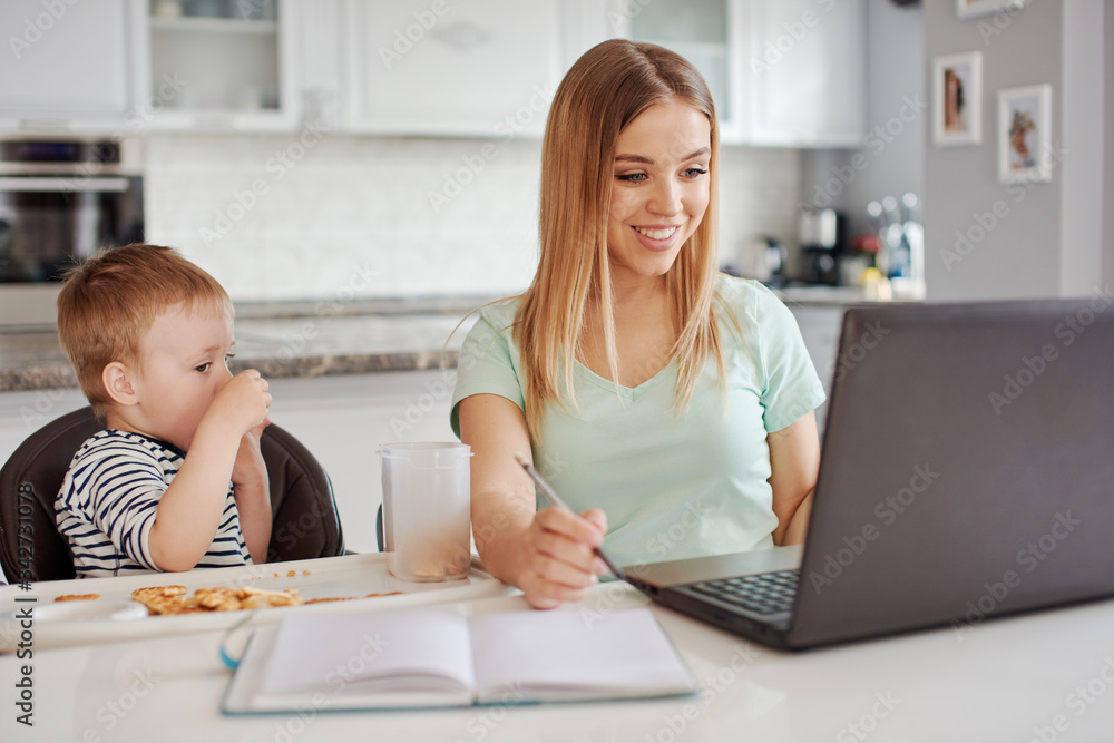 Young woman with child studying on laptop, making notes while feeding baby Young woman with child studying on laptop, making notes while feeding baby
