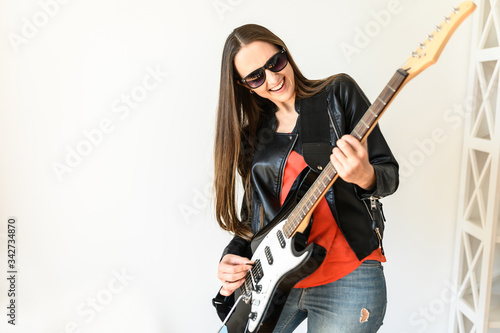A young beautiful woman in sunglasses and in leather jacket is playing an electric guitar indoors, she is smiling happily. Hobby concept, music talent