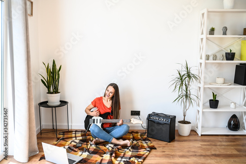 A young beauty girl is learning electric guitar playing with a video tutorial on laptop. She sits on cozy plaid with guitar in hands, combo amp near on the floor photo