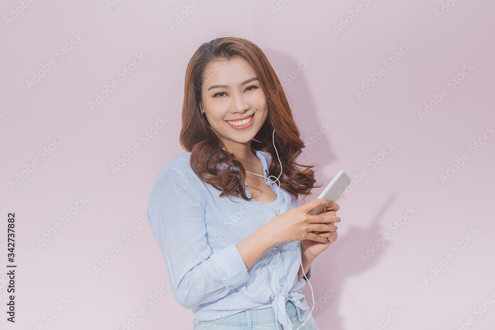 Portrait of a beautiful young woman standing isolated over pink background, listening to music with earphones and mobile phone Portrait of a beautiful young woman standing isolated over pink background, listening to music with earphones and mobile phone