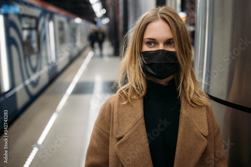 Blonde woman in black medical mask, in coat standing in subway.