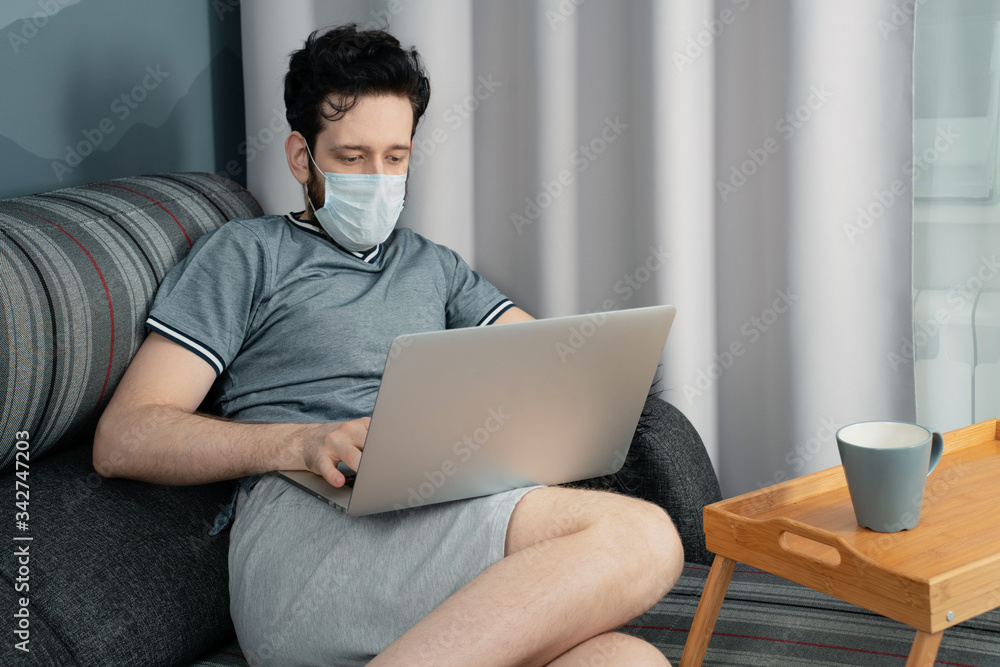 A man in a medical mask is working on a laptop sitting at home on the sofa A man in a medical mask is working on a laptop sitting at home on the sofa