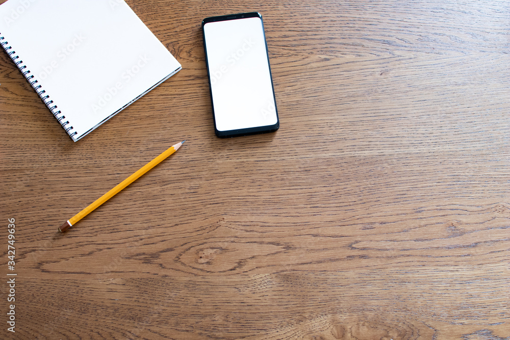 Smartphone with white screen, notebook blank and pencil on wooden table. Working at home during quarantine. Smart working concept with simple tools. Designer and creativity at home Smartphone with white screen, notebook blank and pencil on wooden table. Working at home during quarantine. Smart working concept with simple tools. Designer and creativity at home