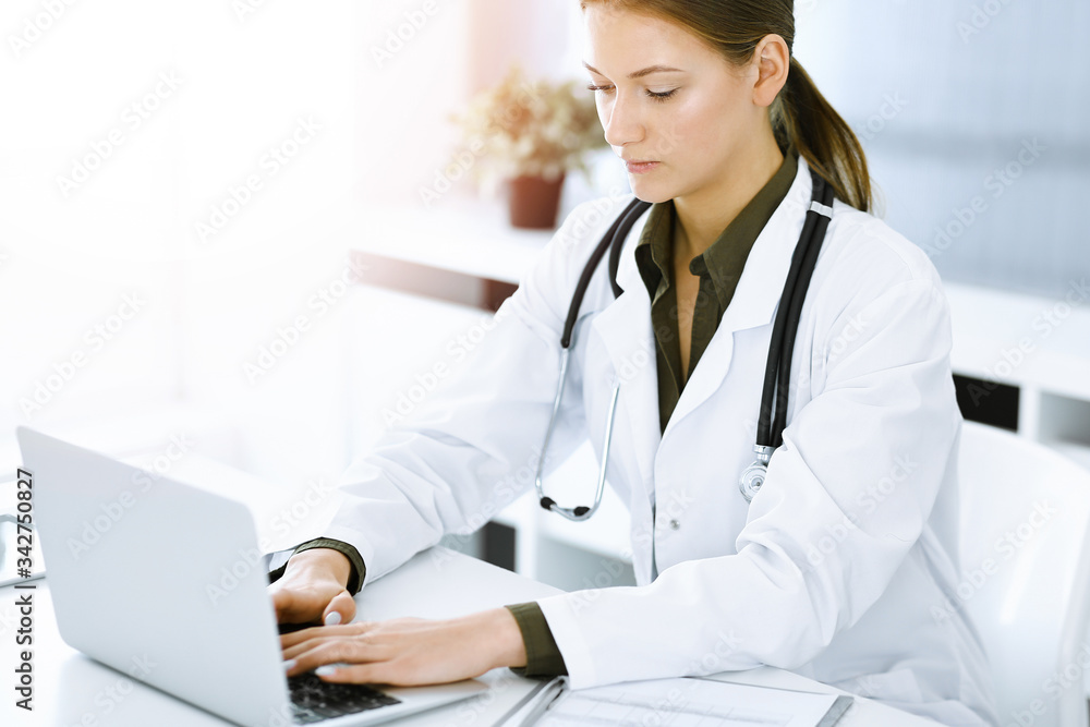 Woman-doctor typing on laptop computer while sitting at the desk in sunny hospital office. Physician at work Woman-doctor typing on laptop computer while sitting at the desk in sunny hospital office. Physician at work