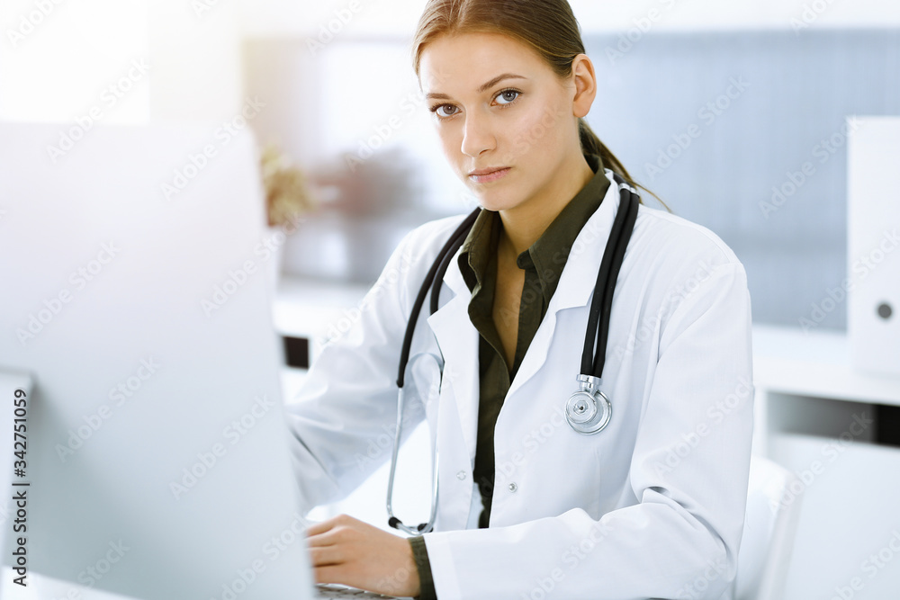 Woman-doctor typing on pc computer while sitting at the desk in sunny hospital office. Data in medicine and healthcare Woman-doctor typing on pc computer while sitting at the desk in sunny hospital office. Data in medicine and healthcare