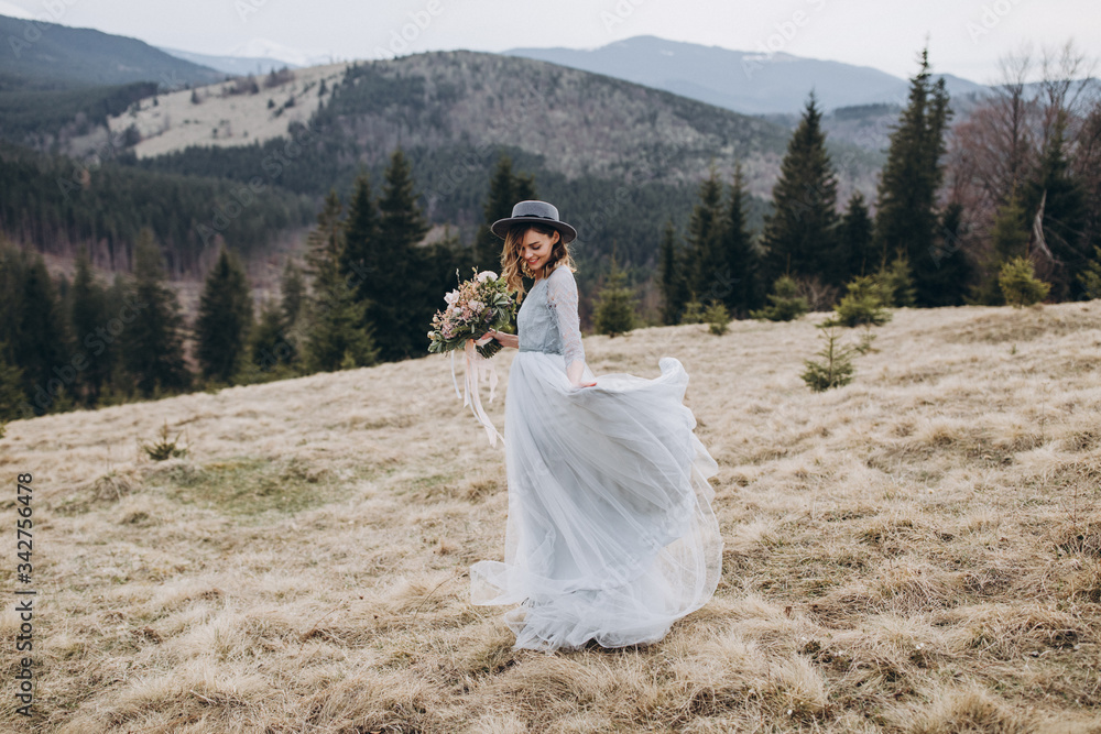 Portrait of the bride. A young girl in a blue gray wedding dress and hat with a bouquet of flowers and greenery in her hands on a background of mountains and a forest at sunset