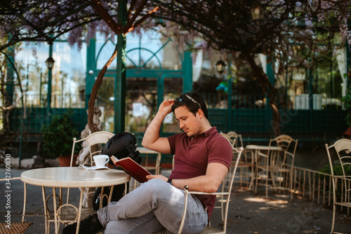A young caucasian man in a cafe garden is reading a book