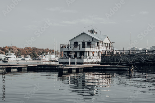 An aesthetic calm view of an authentic boathouse on a river pier. White wooden building with a parked yachts. Reflection on the water. Cloudy sky. Travel and sailing concept. River landscape © Марина Григорьева