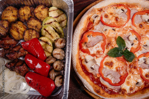 Roasted potatoes with grilled vegetables and meat on baking tray and pizza on wooden background