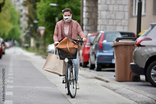Young woman with a white-patterned DIY protective mask driving a bicycle wearing a white shopping bag.