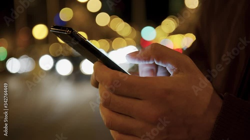 Closeup of male hand typing a cell phone screen and web surfing while standing near a night road. Handheld real time close-up shot.