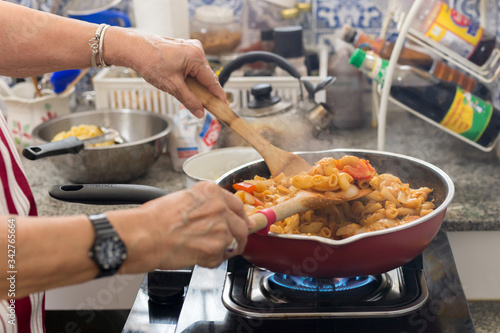 Old woman cooking macaroni pasta in flying pan in the kitchen.