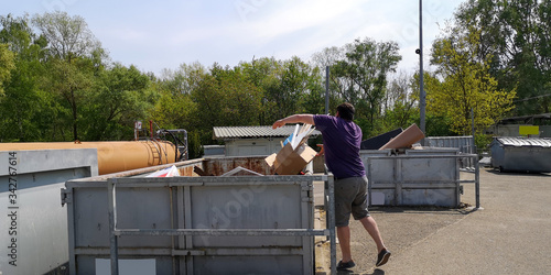 фотографія Plump,dark-haired,middle-aged man throws cardboard box with household waste into huge container at station of collection of large-size garbage in Germany, Europe on sunny day