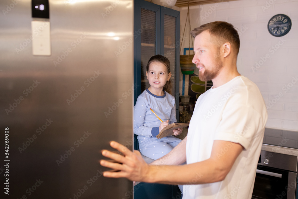 Fototapeta premium Young man opening door of refrigerator while his daughter making shopping list